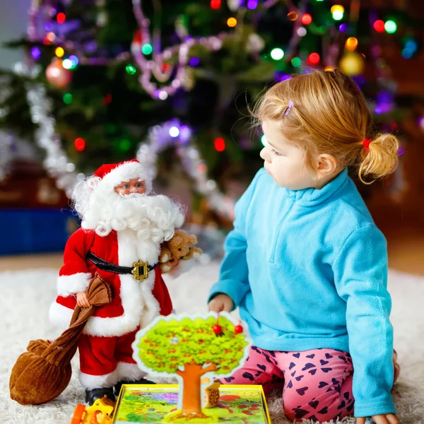 Adorable toddler girl playing with gifts and Christmas Santa Claus toys. Little child having fun with decorated and illuminated Xmas tree with lights on background. Happy healthy funny baby girl. — Stock Photo, Image