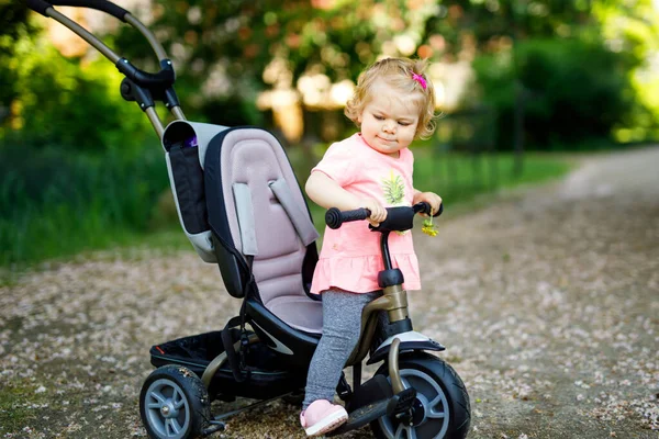 Χαριτωμένο, αξιολάτρευτο κοριτσάκι που κάθεται πάνω στο ποδήλατο ή το τρίκυκλο. Ένα μωράκι που πάει βόλτα με τους γονείς του την ηλιόλουστη μέρα. Ευτυχισμένο υγιές παιδί με πολύχρωμα ρούχα — Φωτογραφία Αρχείου