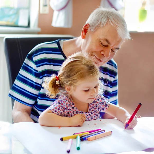 Schattig klein baby peuter meisje en knappe senior grootvader schilderij met kleurrijke potloden thuis. Kleinkind en man hebben plezier samen. Familie en generatie in liefde — Stockfoto