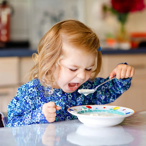 おいしい幼児の女の子は朝食のためにミルクと健康的なシリアルを食べています。キッチンに座って、オート、シリアルを準備するのが楽しいカラフルな服のかわいい幸せな赤ちゃん。家の中で — ストック写真