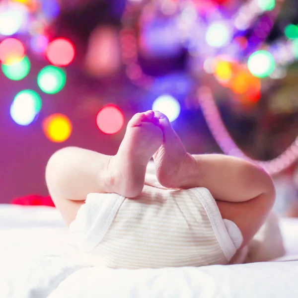 Closup van pasgeboren baby voeten en luier met kerstboom en kleurrijke garland verlichting op de achtergrond tot op de grond met schattige kleine voeten. Close-up. Familie, geboorte, feest concept. — Stockfoto