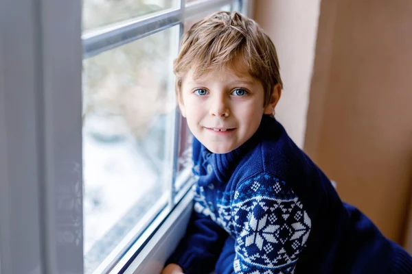 Niño adorable feliz sentado cerca de la ventana y mirando hacia fuera en la nieve en el día de Navidad o la mañana. Sonriente niño sano fascinado observando nevadas y grandes copos de nieve — Foto de Stock