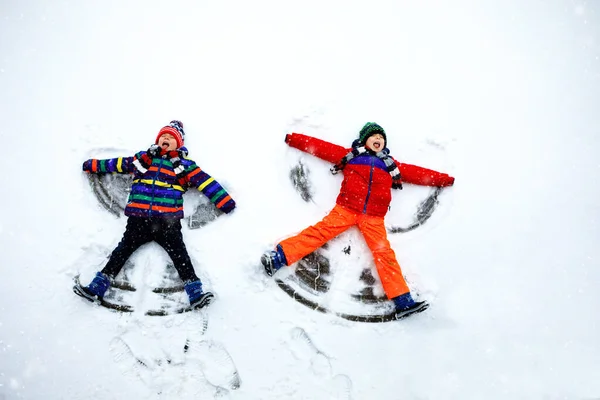 Két kistestvér fiú színes téli ruhában hóangyalt csinál, hóra fektetve. Aktív szabadtéri kikapcsolódás gyerekekkel télen. Boldog testvérek meleg kalap, kesztyű, téli divat. — Stock Fotó