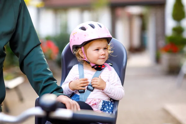 Portrait of little toddler girl with security helmet on the head sitting in bike seat and her father or mother with bicycle. Safe and child protection concept. Family and weekend activity trip.