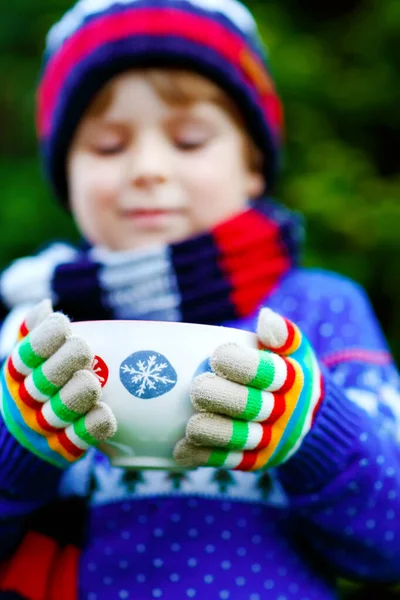 Happy little kid boy drinking hot cocoa and chocolate drink and marshmallows. Funny child in winter sweater, cap, long warm scarf and colorful gloves. On cold winter day during snowfall outdoors — Stock Photo, Image