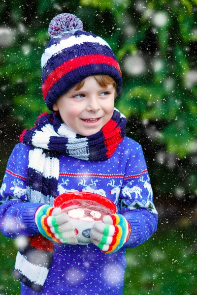 Glücklicher kleiner Junge trinkt heißen Kakao und Schokoladengetränk und Marshmallows. Lustiges Kind in Winterpullover, Mütze, langem warmen Schal und bunten Handschuhen. An kalten Wintertagen bei Schneefall im Freien — Stockfoto