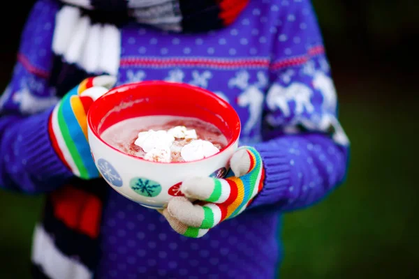 Zbliżenie rąk dziecka trzymającego duży kubek z płatkami śniegu i gorącym kakao i napojem czekoladowym i piankami. Dzieciak w zimowym swetrze, długi ciepły szalik i kolorowe rękawiczki. W chłodny zimowy dzień — Zdjęcie stockowe