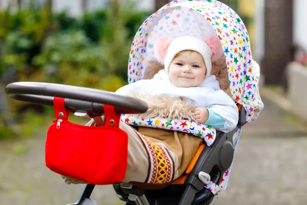 Bonito pequena menina bonita sentada no carrinho de bebê ou carrinho no dia de outono. Criança sã feliz que vai para um passeio no ar puro na roupa quente. Bebê com roupas coloridas e chapéu com bobbles — Fotografia de Stock