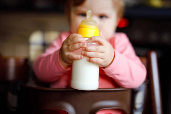 Linda menina adorável segurando mamadeira e bebendo leite fórmula. Primeira comida para bebés. Criança recém-nascida, sentada na cadeira da cozinha doméstica. Bebês saudáveis e conceito de alimentação por mamadeira — Fotografia de Stock