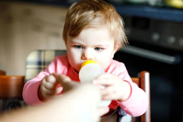 Linda niña adorable sosteniendo biberón y beber leche maternizada. Primera comida para bebés. Niño recién nacido, sentado en la silla de la cocina doméstica. Bebés sanos y concepto de alimentación con biberón — Foto de Stock