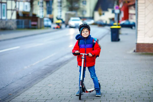 Hermoso niño de escuela montado en scooter camino a la escuela primaria. Niño con casco de seguridad, bolso escolar en otoño lluvioso o mañana fría invierno. Tráfico en la ciudad y escolares —  Fotos de Stock