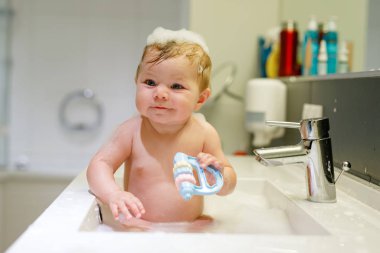 Lavaboda yıkanan ve musluğu tutan sevimli bir bebek. Büyük mavi gözlü, köpükle oynayan ve eğlenen küçük sağlıklı bir kız..