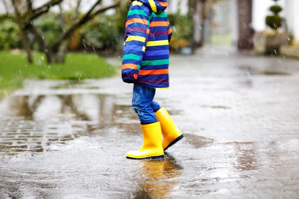 Primo piano del bambino che indossa stivali da pioggia gialli e cammina durante la nevicata, la pioggia e la neve nelle giornate fredde. Bambino in abiti casual moda colorata saltare in una pozzanghera. Divertirsi all'aria aperta — Foto Stock