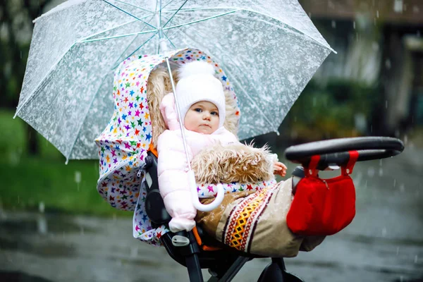 Cute little beautiful baby girl sitting in the pram or stroller on cold day with sleet, rain and snow. Happy smiling child in warm clothes, fashion stylish baby coat. Baby with big umbrella — Stock Photo, Image