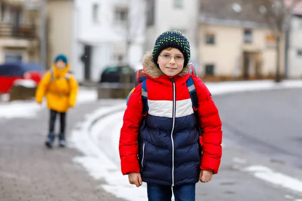 Liten skola unge pojke på elementär klass går till skolan under snöfall. Glada barn och studerande med glasögon ryggsäck i färgglada vinterkläder. schoolkid i gul jacka på bakgrund — Stockfoto