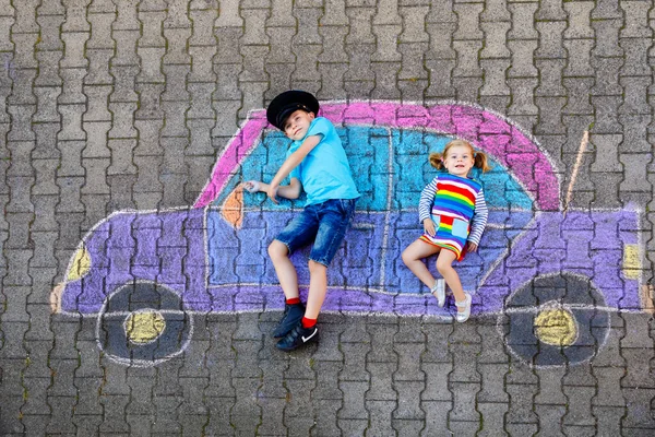 Zwei kleine Kinder, ein kleiner Junge und ein kleines Mädchen amüsieren sich mit einer Autobildzeichnung mit bunten Kreiden auf Asphalt. Geschwister malen auf dem Boden und spielen zusammen. Kreative Freizeit für Kinder — Stockfoto