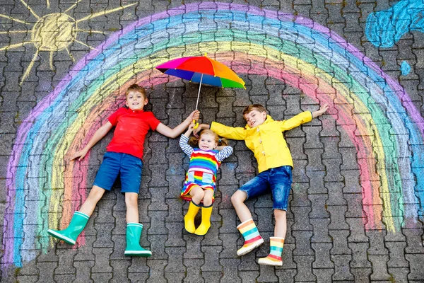 Three little children, two school kids boys and toddler girl having fun with with rainbow picture drawing with colorful chalks on asphalt. Siblings in rubber boots painting on ground playing together. — Stock Photo, Image