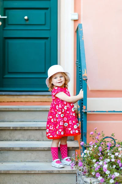 Portrait of beautiful little gorgeus lovely toddler girl in pink summer look clothes, fashion dress, knee socks and hat. Happy healthy baby child posing infront of colorful house. — Stock Photo, Image