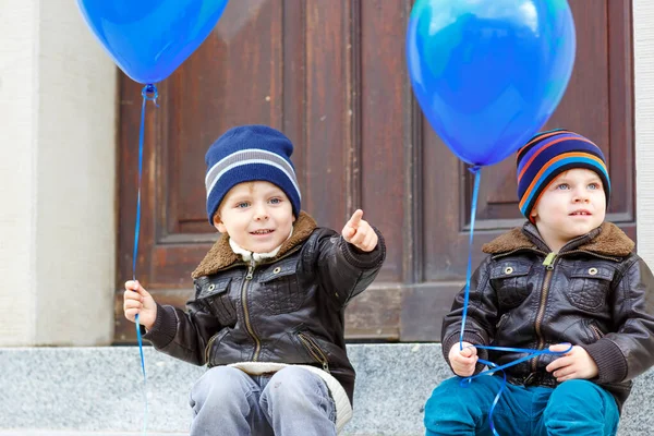 Två barn lite pojkar spelar med blå luft ballonger utomhus. Happy tvillingar och småbarn bröder ler och skrattar tillsammans. Friska barn utanför kall dag i varma kläder. — Stockfoto