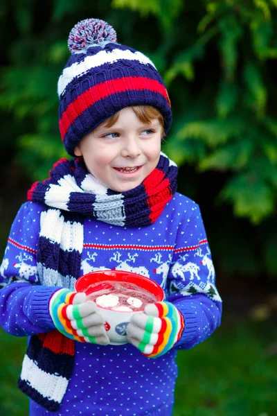 Glücklicher kleiner Junge trinkt heißen Kakao und Schokoladengetränk und Marshmallows. Lustiges Kind in Winterpullover, Mütze, langem warmen Schal und bunten Handschuhen. An kalten Wintertagen bei Schneefall im Freien — Stockfoto