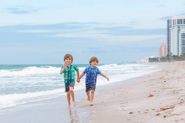 Due ragazzini felici che corrono sulla spiaggia dell'oceano. Bambini simpatici divertenti, fratelli e migliori amici fare vacanze e godersi l'estate nella giornata tempestosa ventosa. Bambini sani sulla spiaggia di Miami, Stati Uniti d'America — Foto Stock