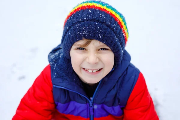 Porträtt av en liten skolpojke i färgglada kläder som leker utomhus under snöfallet. Aktiv fritid med barn på vintern på kalla snöiga dagar. Glad frisk barn att ha kul och leka med snö. — Stockfoto