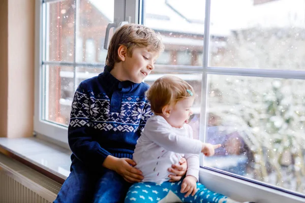 Glad bedårande pojke och söt liten flicka sitter nära fönstret och tittar ut på snö på juldagen eller morgonen. Leende barn, syskon, lillasyster och bror tittar på vintern snöfall. — Stockfoto