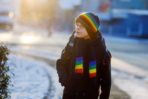 Niño de la escuela de primaria caminando a la escuela durante las nevadas. Feliz niño sano con gafas divirtiéndose y jugando con la nieve. Con mochila o mochila en ropa de invierno colorida. — Foto de Stock