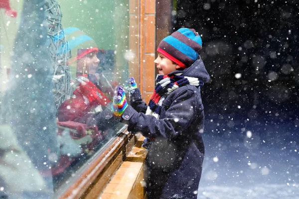 Słodki mały zdrowy chłopiec ze szkoły na rynku świątecznym. Zabawne szczęśliwe dziecko w modzie zimowe ubrania co zakupy okno ozdobione prezentami, choinka. Śnieg pada, śnieg pada — Zdjęcie stockowe