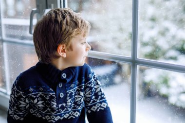 Mutlu sevimli çocuk pencerenin yanında oturuyor ve Noel sabahı ya da sabahleyin dışarıda karları seyrediyor. Gülümseyen sağlıklı çocuk, karları ve büyük kar tanelerini izlerken büyülenir.