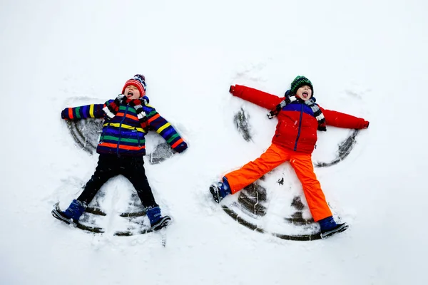 İki küçük kardeş, renkli kışlık giysiler içinde kar meleği yapıp kara uzanan çocuk. Kışın çocuklarla açık hava eğlencesi. Sıcak şapkalı, eldivenli, kış modasında mutlu kardeşler.. — Stok fotoğraf