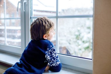 Mutlu sevimli çocuk pencerenin yanında oturuyor ve Noel sabahı ya da sabahleyin dışarıda karları seyrediyor. Gülümseyen sağlıklı çocuk, karları ve büyük kar tanelerini izlerken büyülenir.