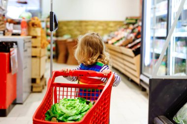 Süpermarkette alışveriş arabasını iten tatlı kız. Küçük çocuk meyve alıyor. Çocuk bakkal alışverişi. Tramvayı olan sevimli bebek marketten taze sebze seçiyor..