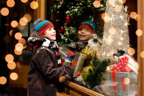 Menino da escola saudável bonito pouco no mercado de Natal. Criança feliz engraçada na moda roupas de inverno fazendo compras de janela decoradas com presentes, árvore de xmas. Neve a cair, neve a cair — Fotografia de Stock