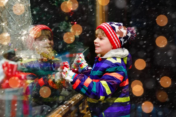 Słodki mały zdrowy chłopiec ze szkoły na rynku świątecznym. Zabawne szczęśliwe dziecko w modzie zimowe ubrania co zakupy okno ozdobione prezentami, choinka. Śnieg pada, śnieg pada — Zdjęcie stockowe