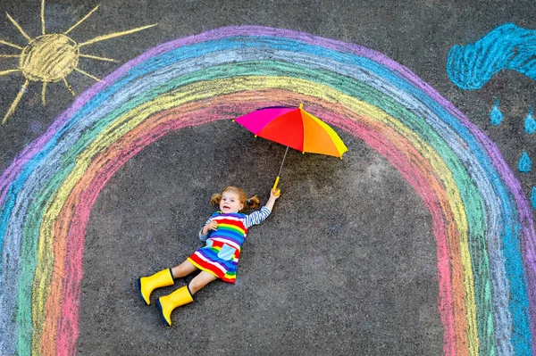 Menina pequena feliz em botas de borracha com sol arco-íris e nuvens com chuva pintada com giz colorido no chão ou asfalto no verão. Criança bonito com guarda-chuva se divertindo. lazer criativo — Fotografia de Stock