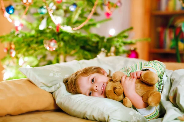 Pequeño niño rubio lindo durmiendo bajo el árbol de Navidad y soñando con Santa en casa, en el interior. Fiesta cristiana tradicional. Niño feliz esperando regalos en Navidad. Acogedora luz suave — Foto de Stock