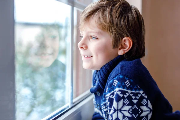 Menino adorável feliz sentado perto da janela e olhando para fora na neve no dia de Natal ou de manhã. Sorrindo criança saudável fascinado observando a queda de neve e grandes flocos de neve — Fotografia de Stock