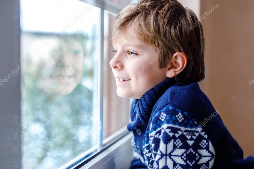Happy adorable kid boy sitting near window and looking outside on snow on Christmas day or morning. Smiling healthy child fascinated observing snowfall and big snowflakes