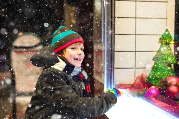 Cute little healthy school kid boy on Christmas market. Funny happy child in fashion winter clothes making window shopping decorated with gifts, xmas tree. Snow falling down, snowfall — Stock Photo, Image