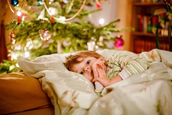 Little cute blond boy sleeping under Christmas tree and dreaming of Santa at home, indoors. Traditional Christian festival. Happy kid child waiting for gifts on xmas. Cozy soft light — Stock Photo, Image