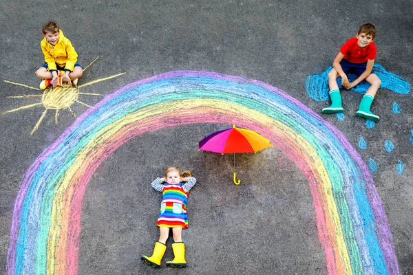 Trois petits enfants, deux écoliers garçons et fillette s'amusent avec dessin d'image arc-en-ciel avec des craies colorées sur asphalte. Frères et sœurs en bottes en caoutchouc peignant sur le sol en jouant ensemble . — Photo