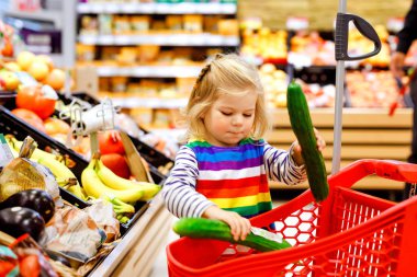 Süpermarkette alışveriş arabasını iten tatlı kız. Küçük çocuk meyve alıyor. Çocuk bakkal alışverişi. Tramvayı olan sevimli bebek marketten taze sebze seçiyor..