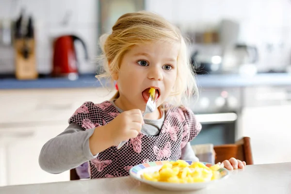 Schöne Kleinkind Mädchen essen gesunde Bratkartoffeln zum Mittagessen. Nettes glückliches Baby in bunten Kleidern, das in der Küche des Hauses, der Kita oder des Kinderzimmers sitzt. Kind isst Gemüse. — Stockfoto