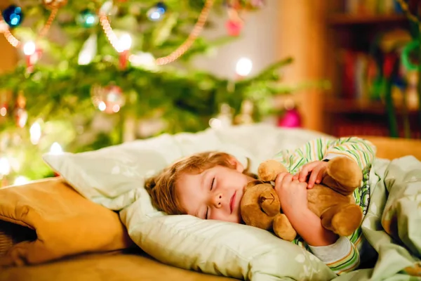 Pequeño niño rubio lindo durmiendo bajo el árbol de Navidad y soñando con Santa en casa, en el interior. Fiesta cristiana tradicional. Niño feliz esperando regalos en Navidad. Acogedora luz suave — Foto de Stock