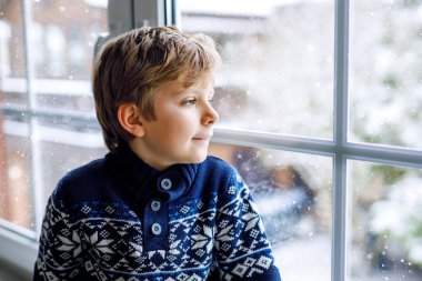 Mutlu sevimli çocuk pencerenin yanında oturuyor ve Noel sabahı ya da sabahleyin dışarıda karları seyrediyor. Gülümseyen sağlıklı çocuk, karları ve büyük kar tanelerini izlerken büyülenir.