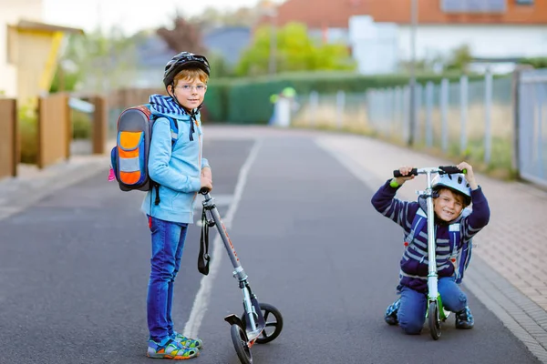 Two school kid boys in safety helmet riding with scooter in the city with backpack on sunny day. Happy children in colorful clothes biking on way to school. — Stock Photo, Image