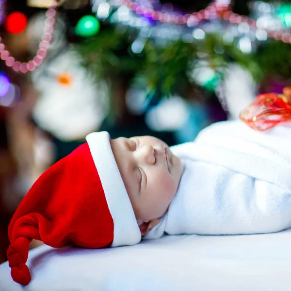 Bebé recién nacido de una semana en sombrero de Santa envuelto en manta cerca del árbol de Navidad con luces de guirnalda de colores en el fondo. Primer plano de un niño lindo, pequeño bebé durmiendo. Familia, Navidad, nacimiento, nueva vida . — Foto de Stock