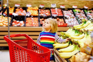 Süpermarkette alışveriş arabasını iten tatlı kız. Küçük çocuk meyve alıyor. Çocuk bakkal alışverişi. Tramvayı olan sevimli bebek marketten taze sebze seçiyor..