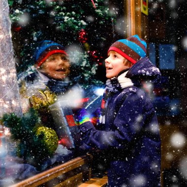 Noel pazarındaki şirin, sağlıklı okul çocuğu. Moda kışı kıyafetleri içinde, hediyelerle süslenmiş vitrin alışverişi yapan neşeli çocuk, xmas ağacı. Kar yağıyor, kar yağıyor.